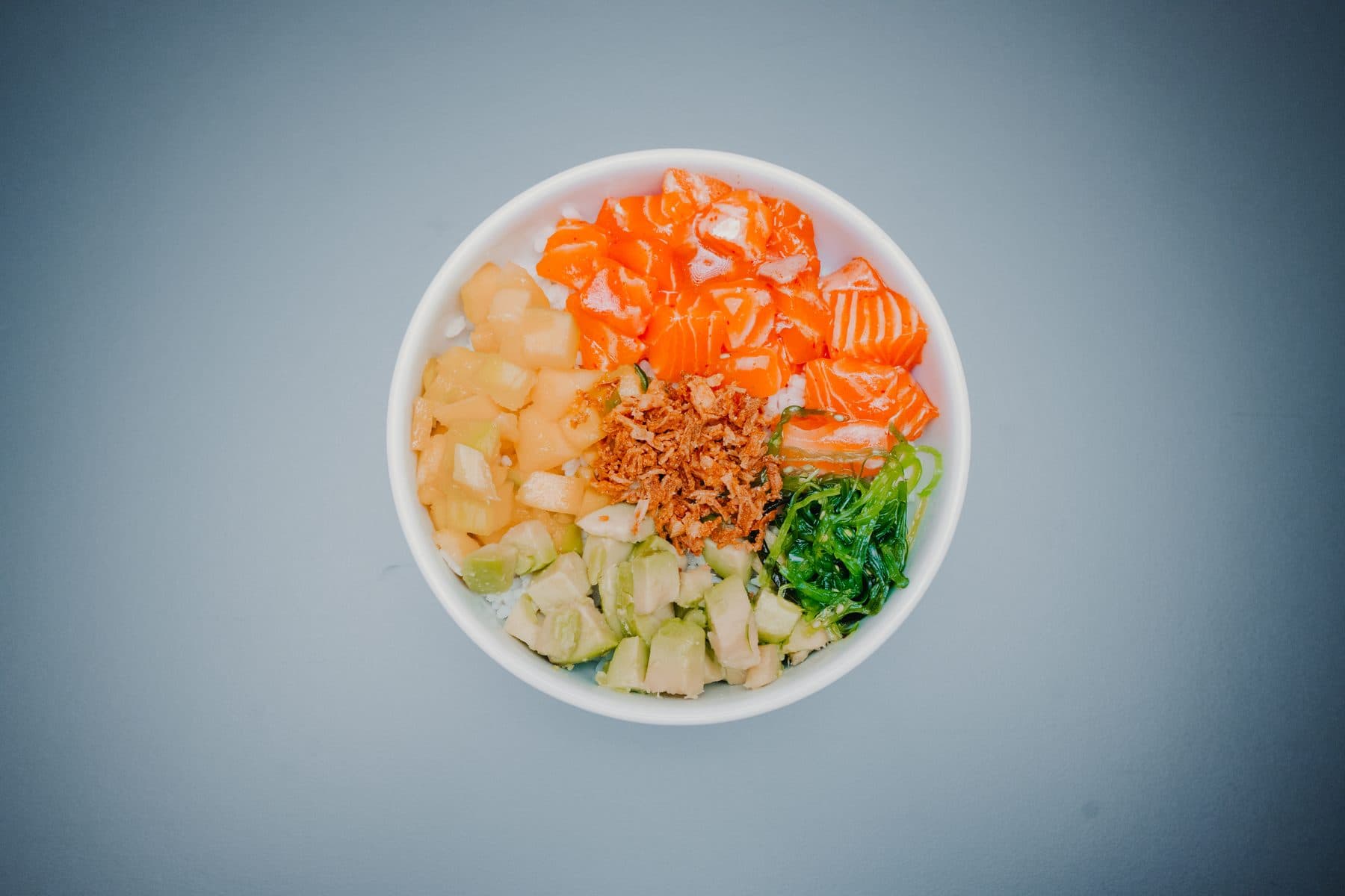 Taça de poke de salmão vista de cima, com algas, cebola frita e outros ingredientes em cubos