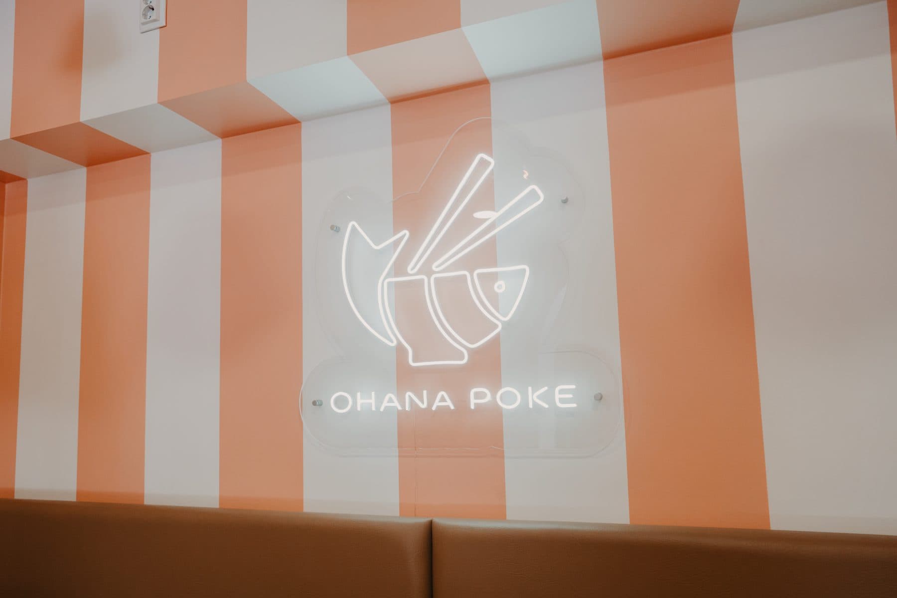 Néon com o logótipo do Ohana Poke numa parede às riscas brancas e cor de salmão, por cima de um banco corrido