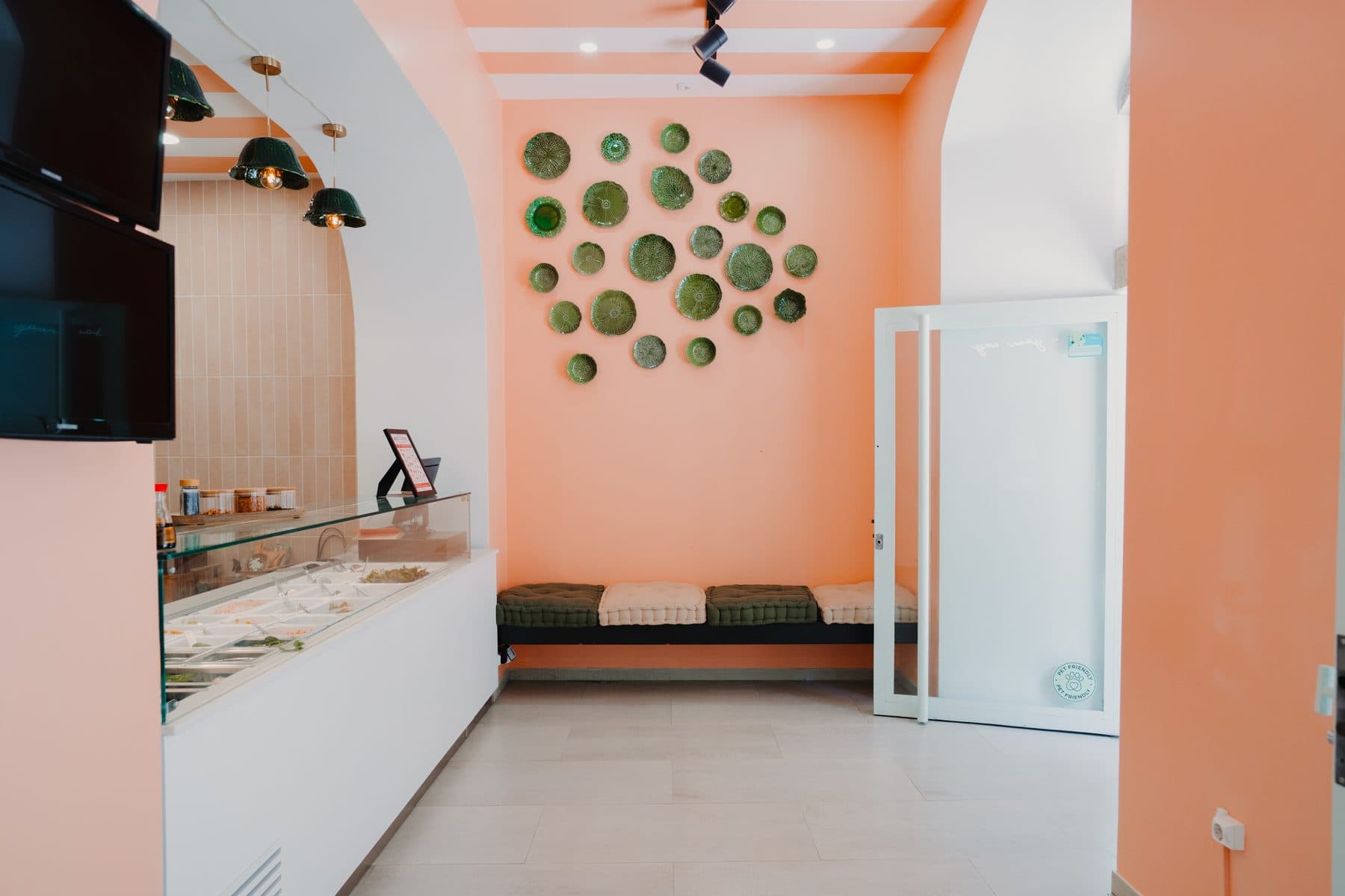 Sala do Ohana Poke com pratos verdes na parede cor de salmão, o balcão à esquerda e um banco com almofadas ao fundo