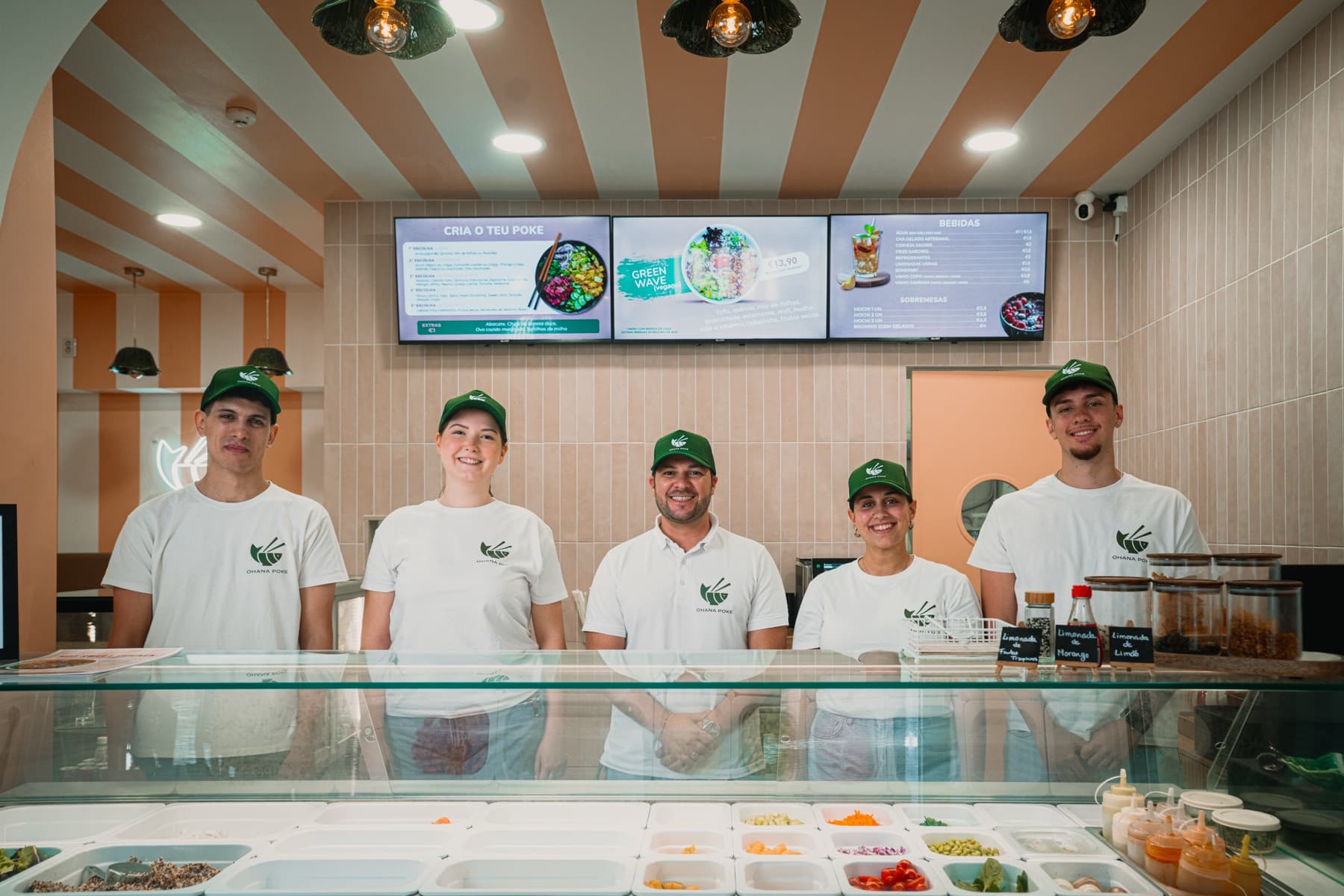 A equipa do Ohana Poke, cinco pessoas de t-shirt branca e boné verde, a sorrir atrás do balcão dos ingredientes