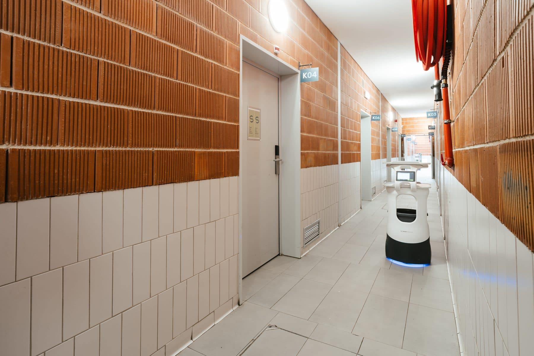 Robô de entregas no corredor das cozinhas, entre a parede de tijolo cor de laranja e as portas numeradas