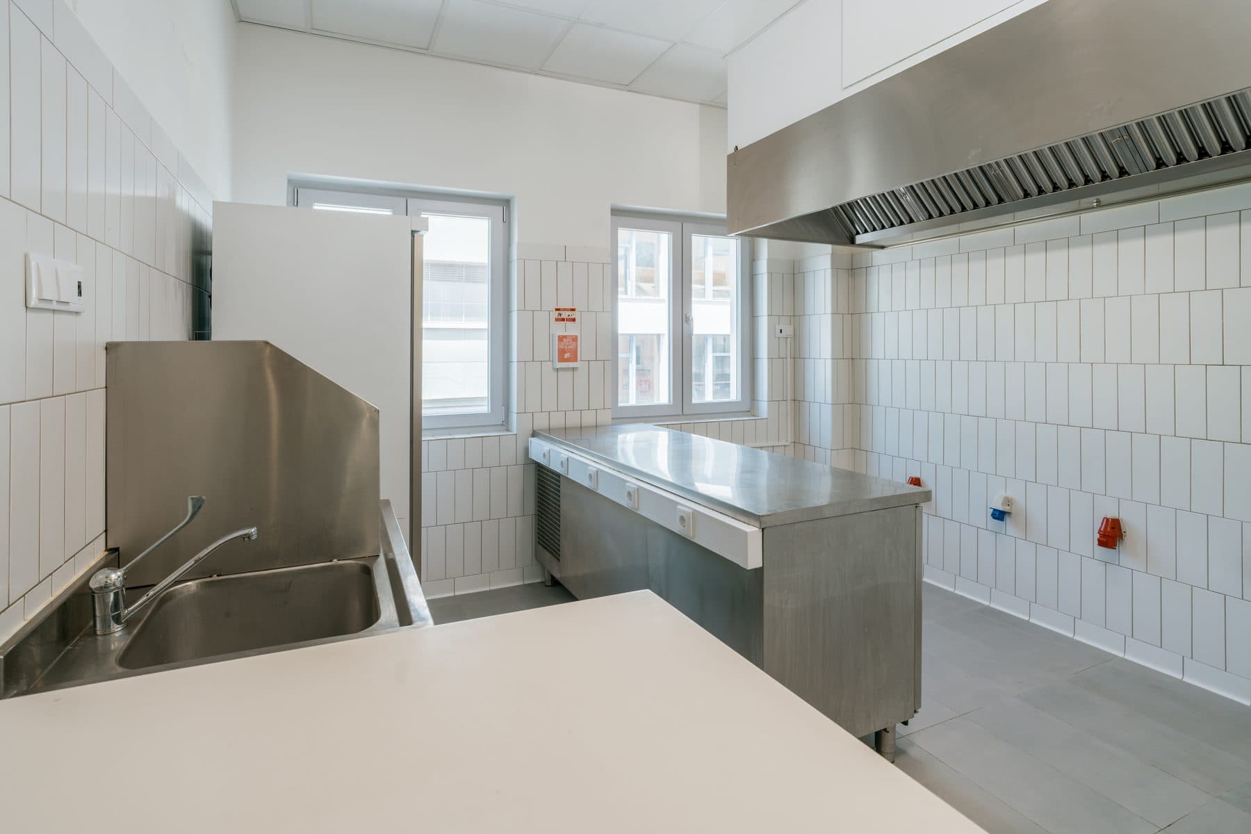 Cozinha vazia, com bancada e lava-loiça em inox, janelas e o exaustor por cima