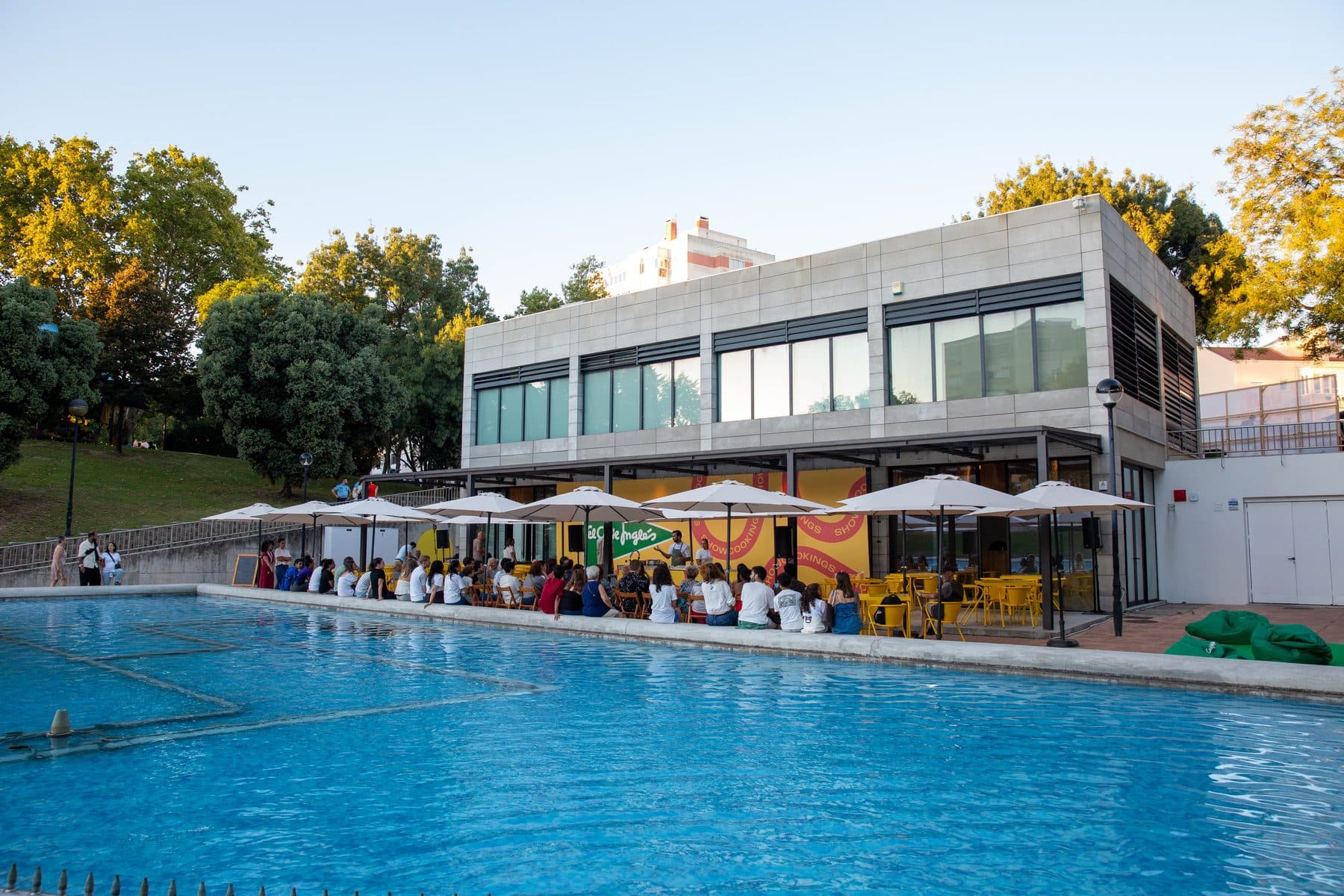 Piscina com a esplanada do festival ao fundo, com guarda-sóis e pessoas sentadas às mesas