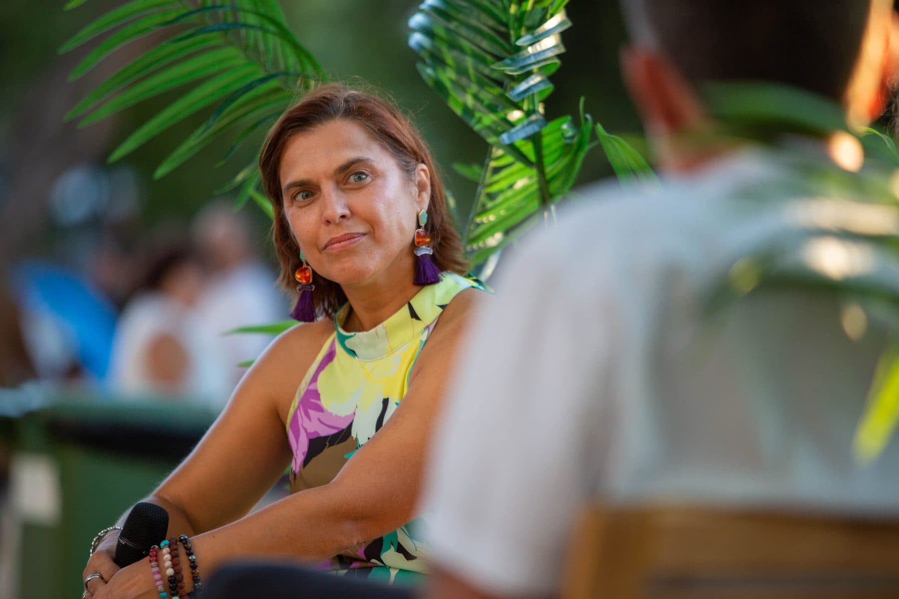 Mulher de microfone na mão, sentada entre plantas, a ouvir numa conversa do festival