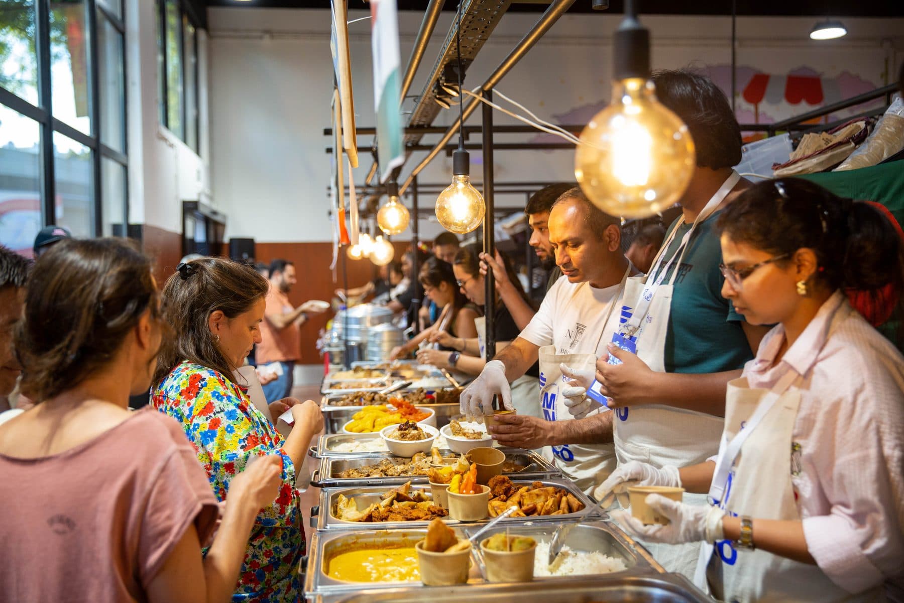 Visitantes a escolher comida numa banca, servidos por uma equipa de luvas atrás dos tabuleiros