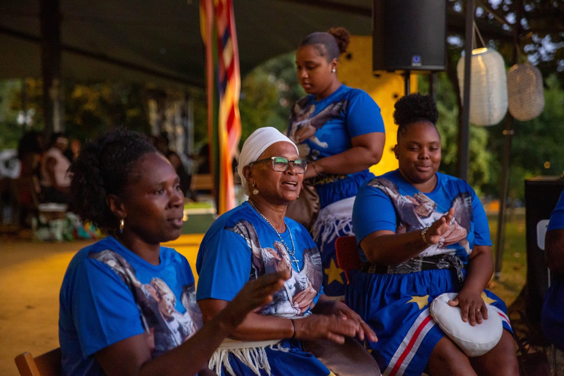 Mulheres de t-shirt azul sentadas a cantar, uma delas com uma almofada branca ao colo