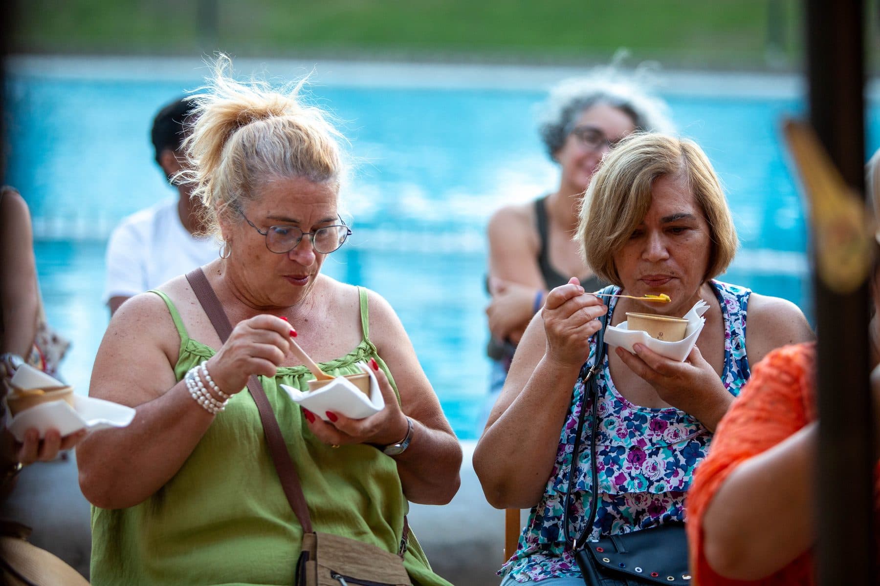 Duas mulheres a comer de copos de papel, sentadas junto à piscina