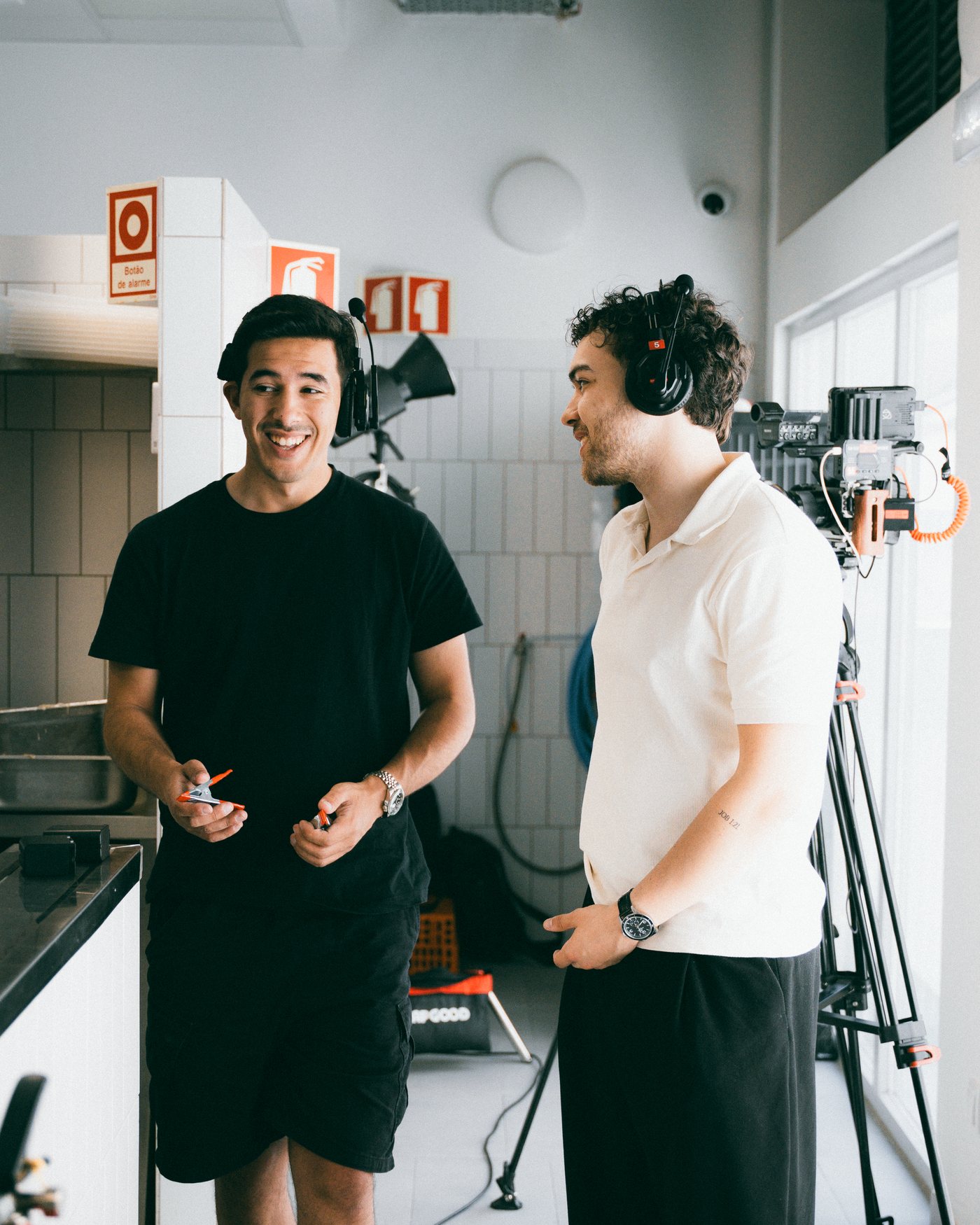 Dois elementos da equipa, de auscultadores, a conversar numa cozinha ao lado da câmara no tripé